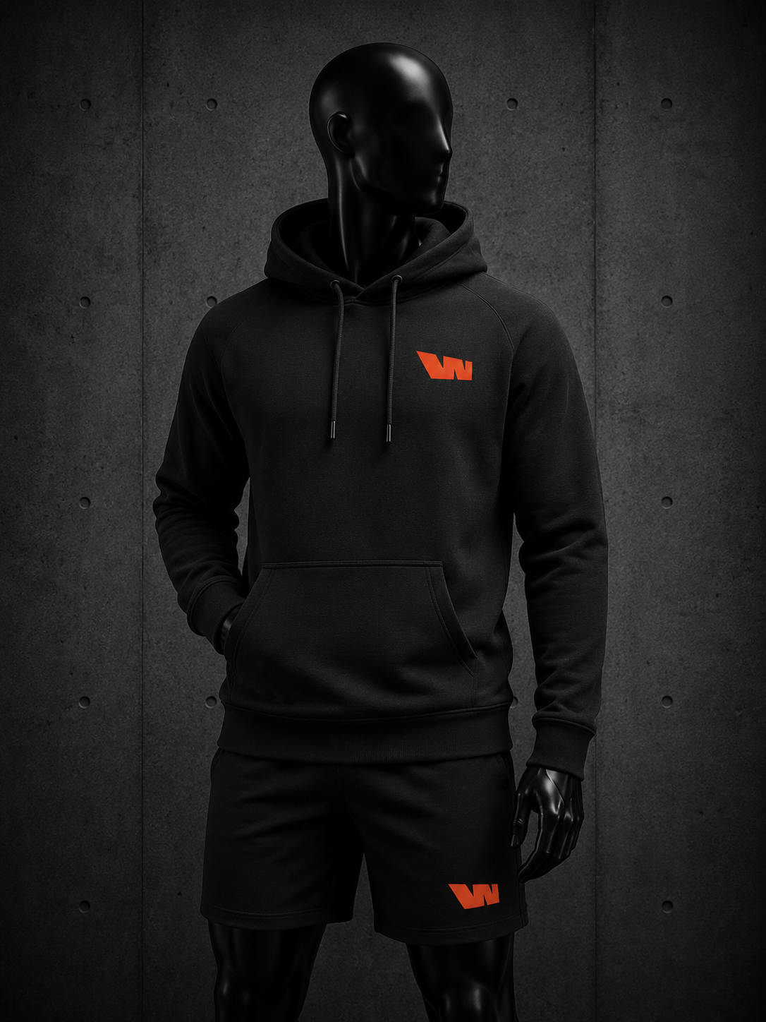A black hoodie and matching black shorts set displayed on a glossy black mannequin against a dark textured wall. The hoodie features a small orange logo on the left chest, and the shorts feature a matching orange logo on the left thigh.